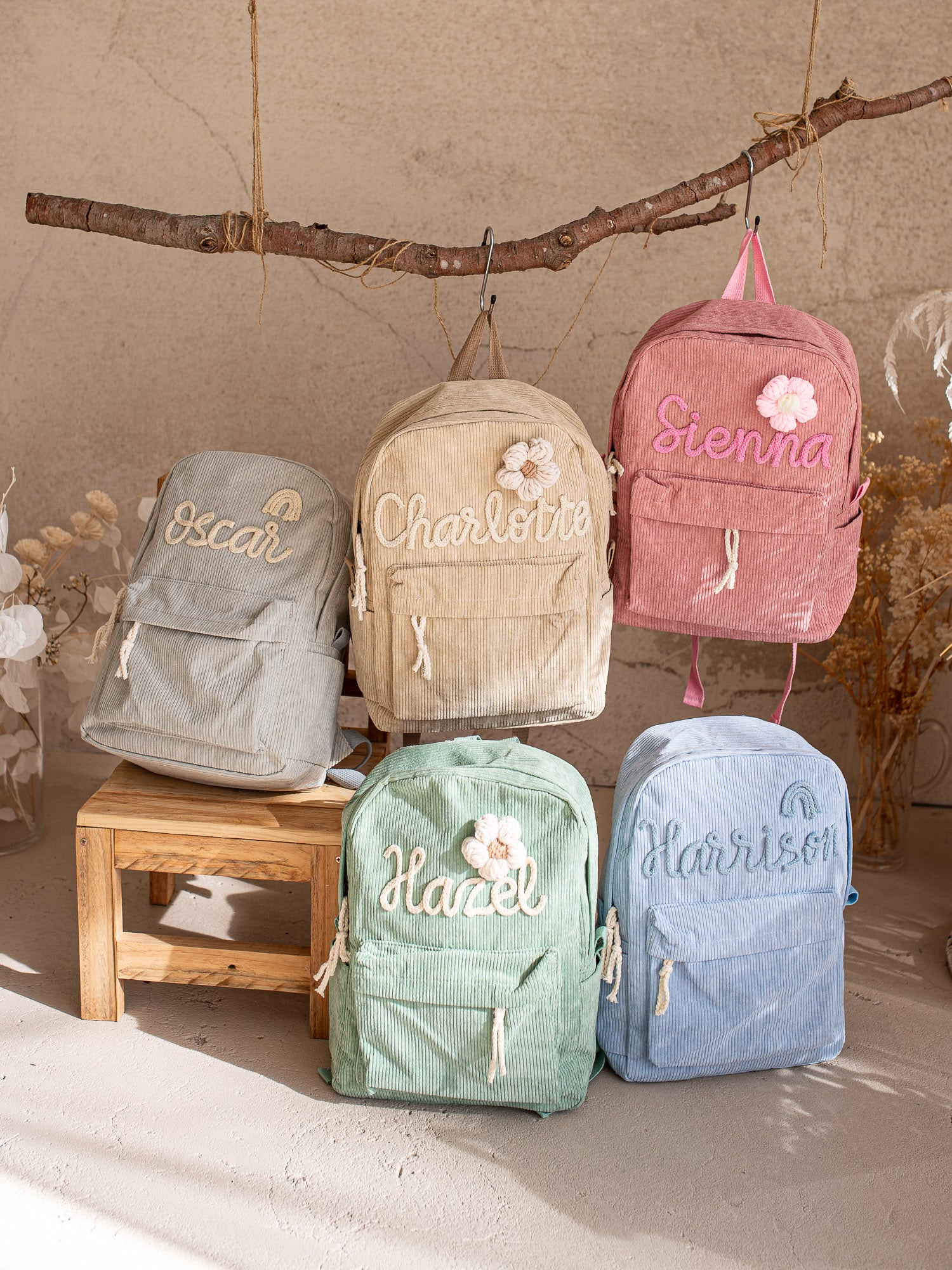 Five corduroy backpacks in grey, beige, pink, green and blue, some hanging from a branch and others on a stool