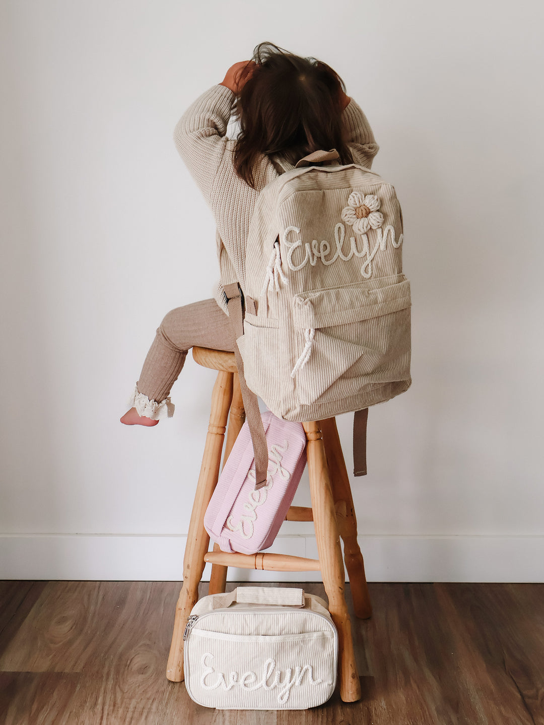 Beige backpack embroidered Evelyn worn by a girl on a stool, matching lunch bag and pink pouch on the floor beneath