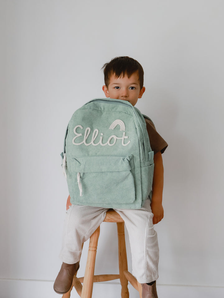 Sage green corduroy backpack embroidered with the name Elliot and a small rainbow, held by a boy sitting on a wooden stool