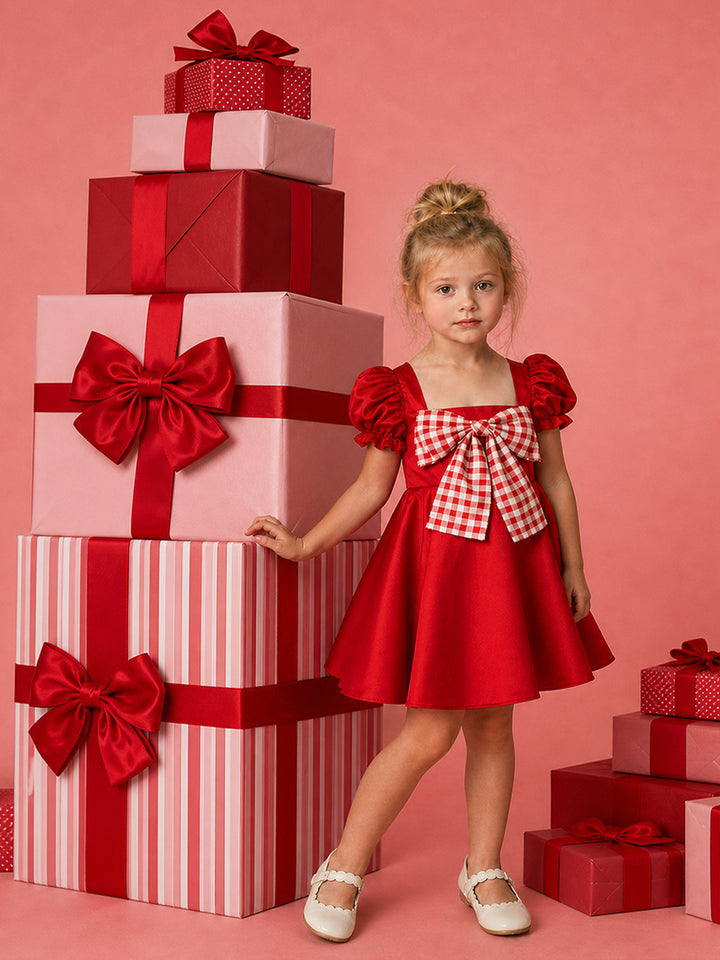 Cherry on Top red satin Christmas dress with puff sleeves and a red gingham bow, standing beside a stack of gift boxes