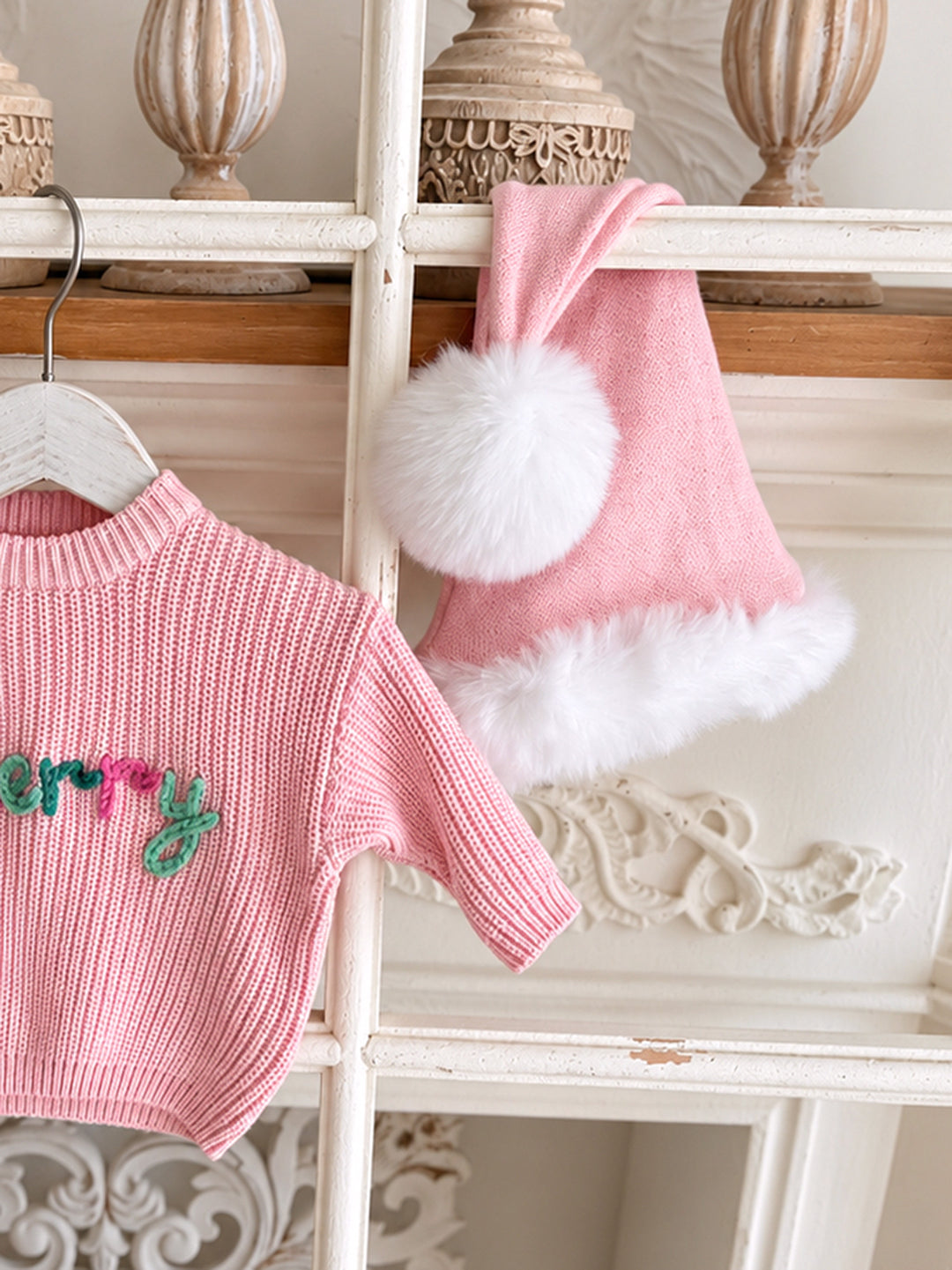 Baby pink knitted Santa hat with white fluffy trim and pom-pom, close up beside a pink Merry knit sweater
