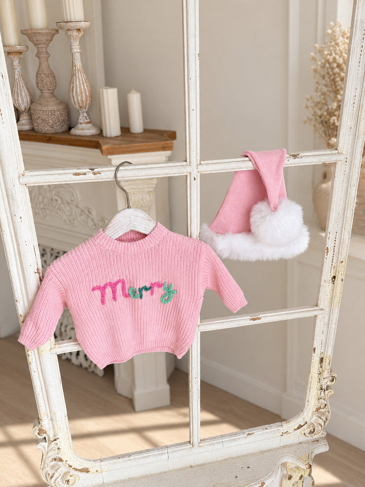Baby pink knitted Santa hat with white fluffy trim and pom-pom, hanging on a vintage window frame beside a pink Merry knit sweater