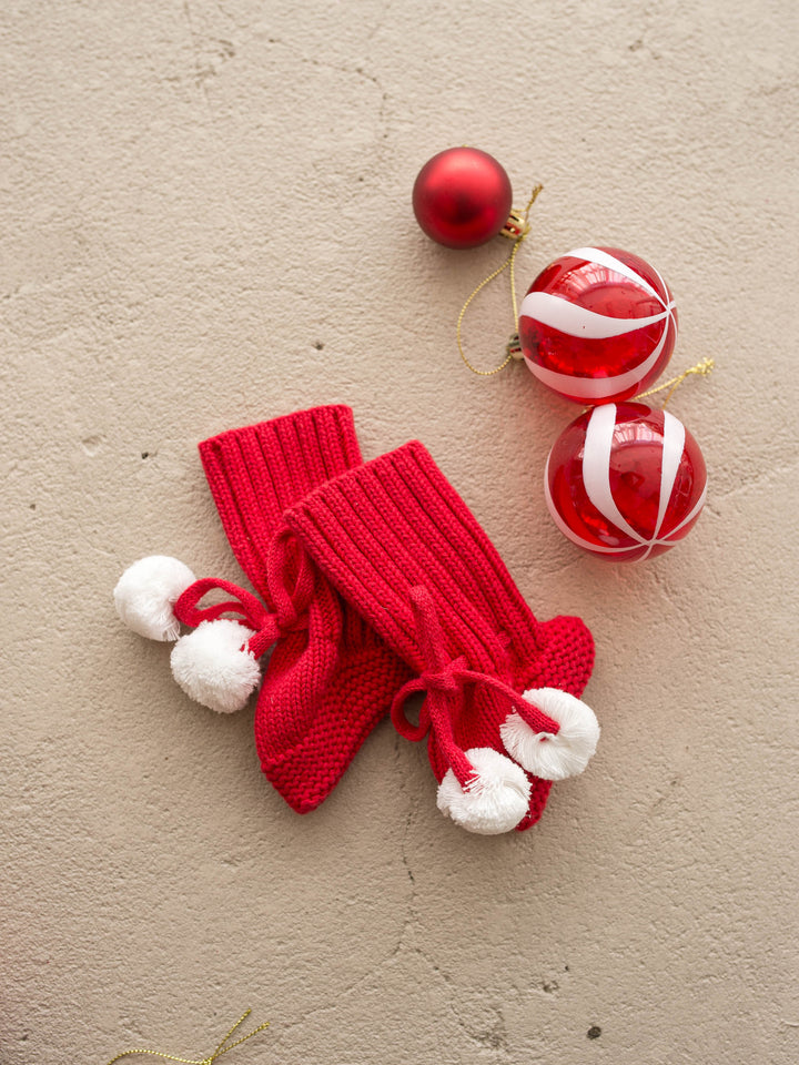 Santa Baby Knitted Booties in red cotton knit with white pom poms, the pair laid flat with red and white baubles - Before & Ever Kids