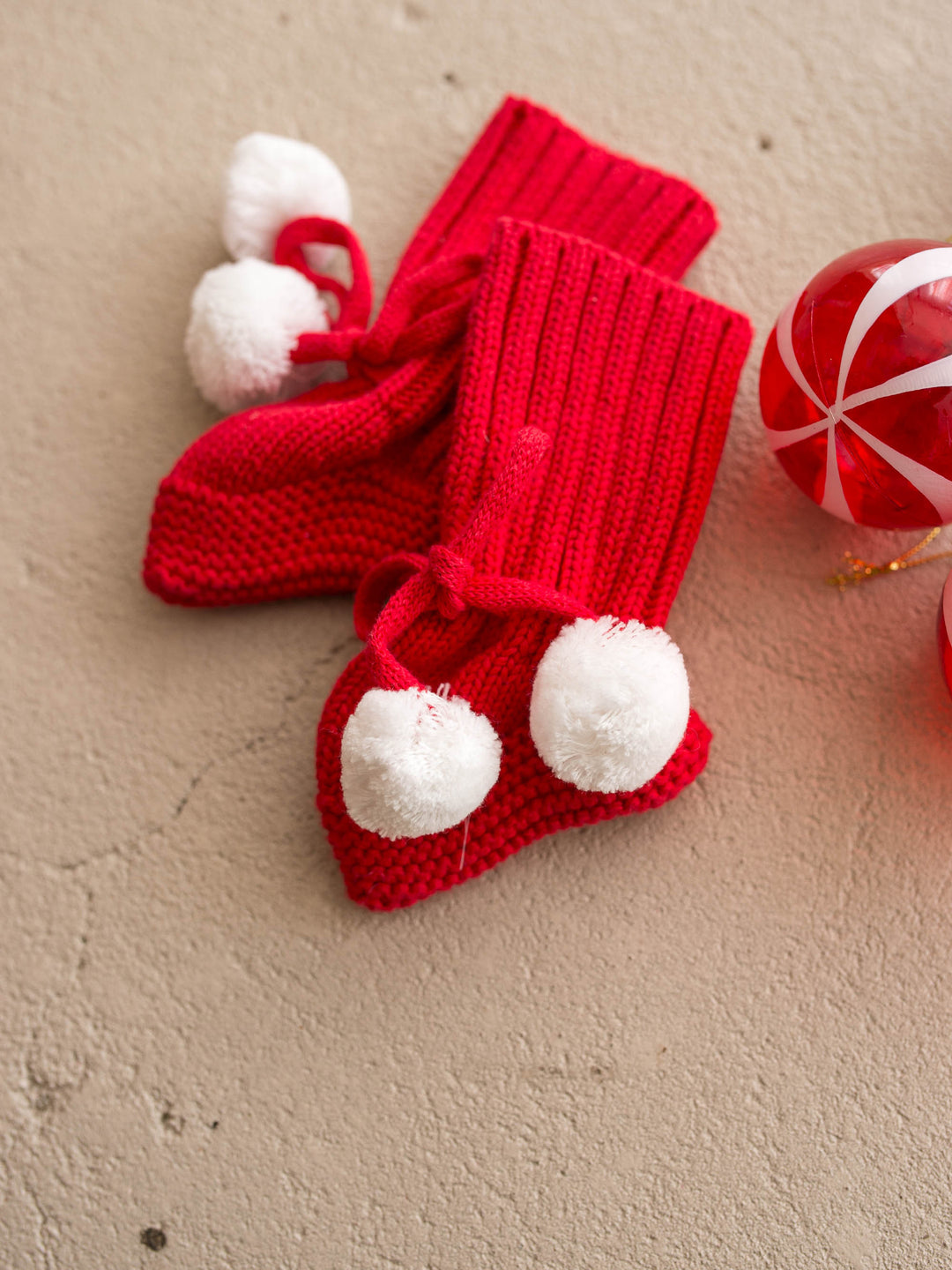 Santa Baby Knitted Booties in red cotton knit with white pom poms, the pair flat with a red bauble - Before & Ever Kids