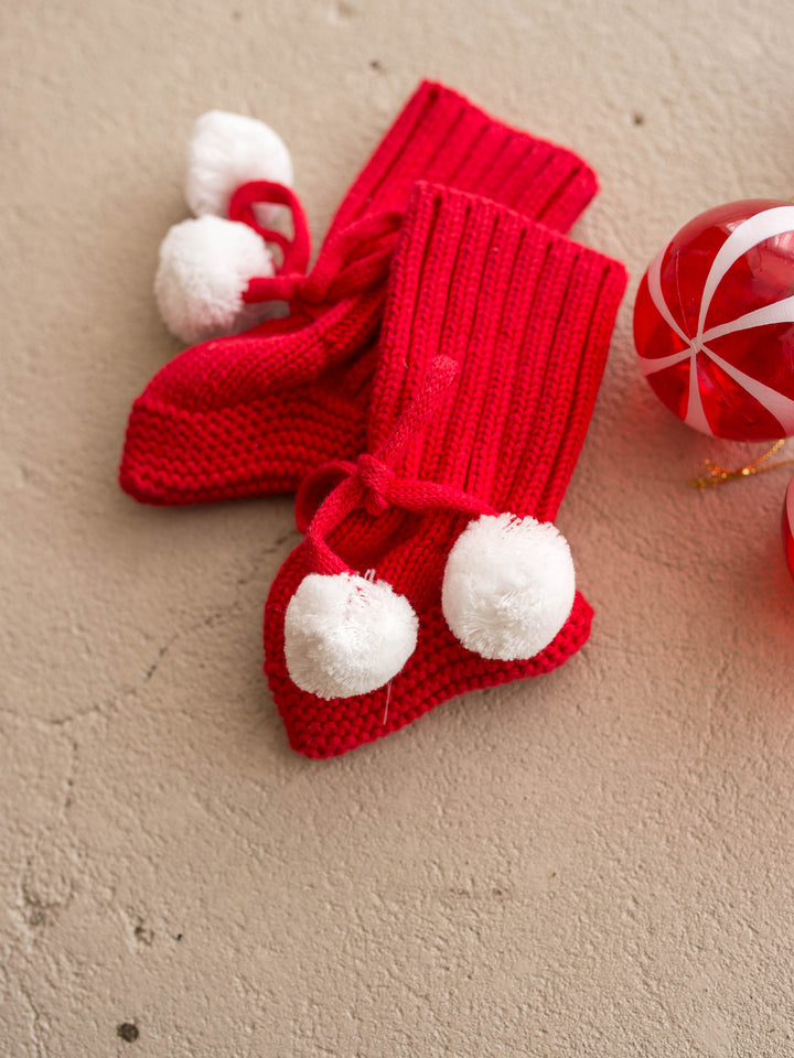 Santa Baby Knitted Booties in red cotton knit with white pom poms, the pair flat with a red bauble - Before & Ever Kids