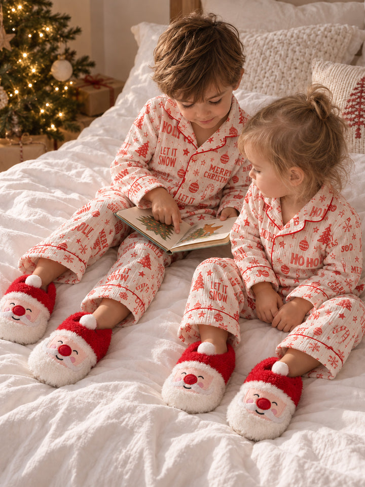 Santa Baby Slippers red plush Santa face slippers worn by two children reading a book on a bed at Christmas - Before & Ever Kids