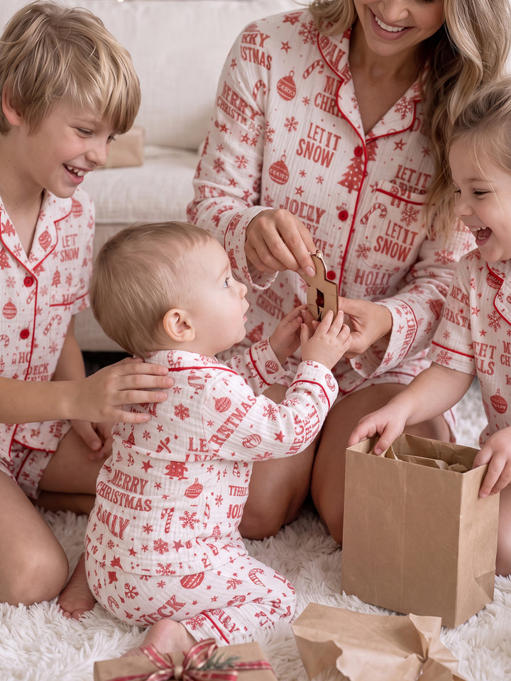 Twas the Night family Christmas pyjamas, white cotton muslin with a red Christmas print, boy, mum, girl and baby laughing over a gift bag - Before & Ever Kids