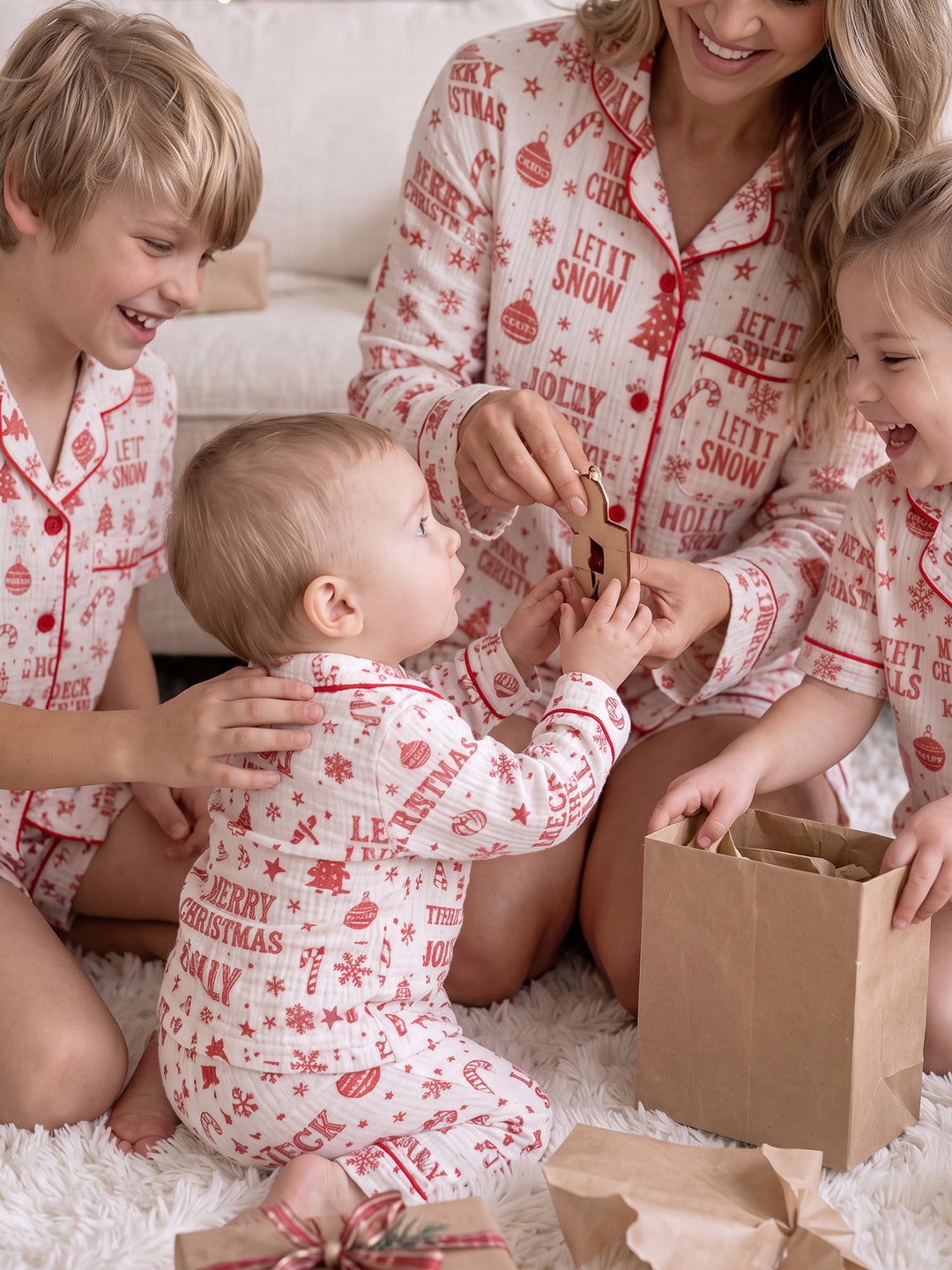 Twas the Night family Christmas pyjamas, white cotton muslin with a red Christmas print, boy, mum, girl and baby laughing over a gift bag - Before & Ever Kids