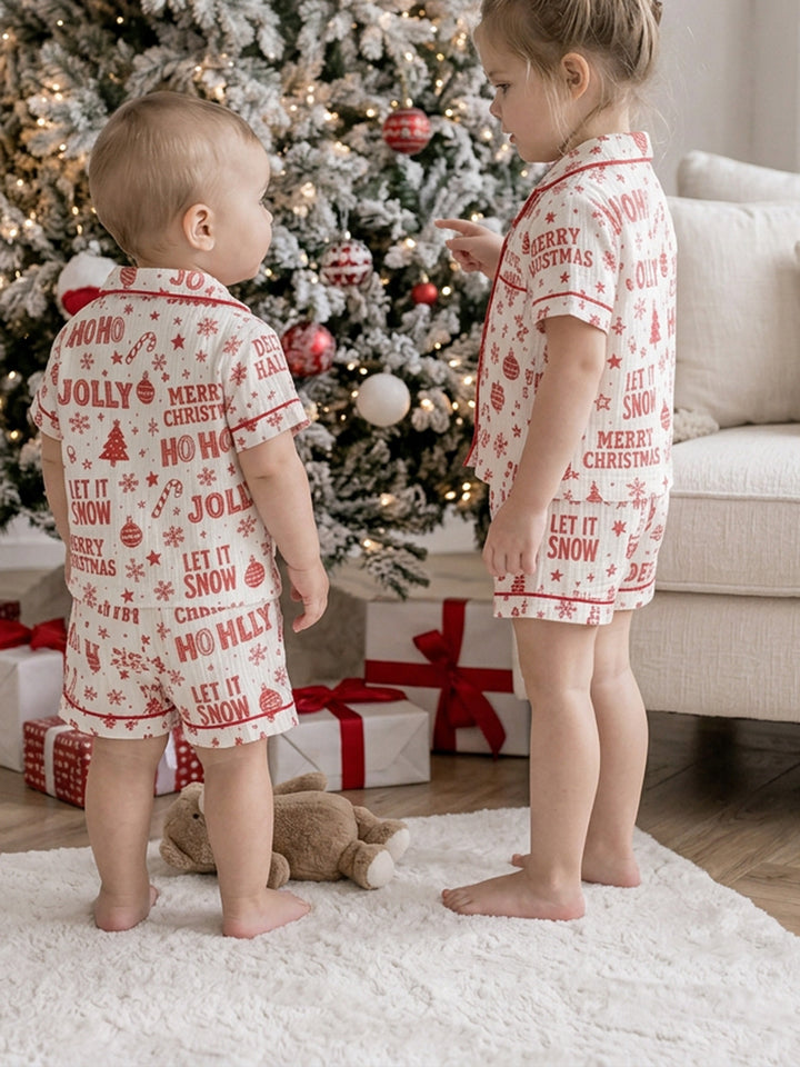 Twas the Night family Christmas pyjamas, white cotton muslin with a red Christmas print, baby and the girl in short sleeve shirts and shorts standing by the tree with a teddy - Before & Ever Kids