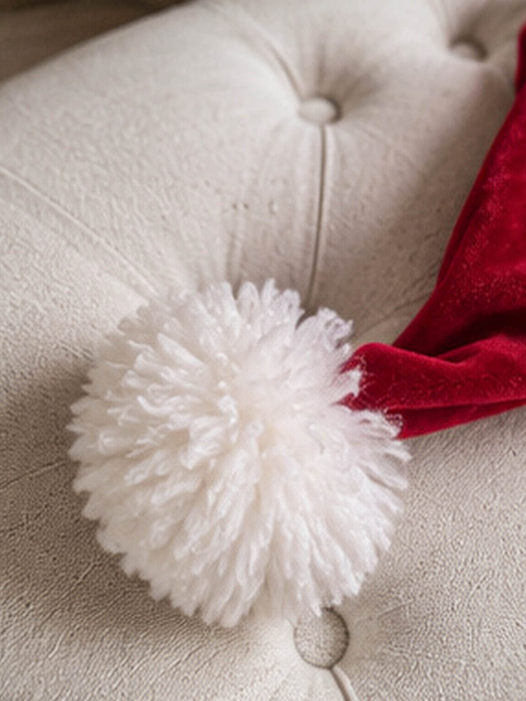 Velvet Santa Hat in deep red, close up of the pom-pom resting on a cream armchair - Before & Ever Kids
